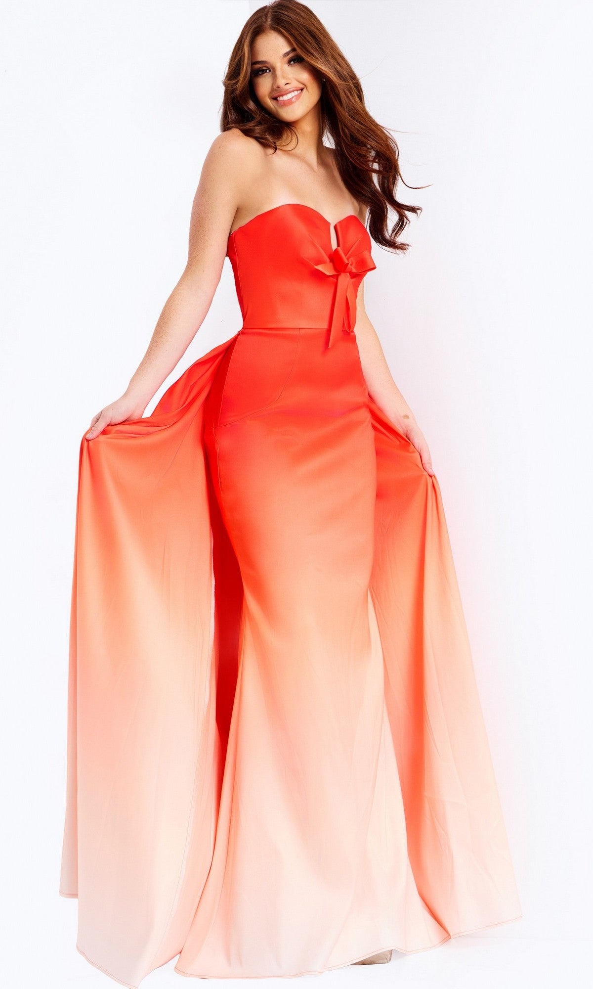 A woman poses while holding the sides of her skirt, wearing the Jovani 47554 Long Prom Dress—an ombre strapless gown with a bow detail at the waist.